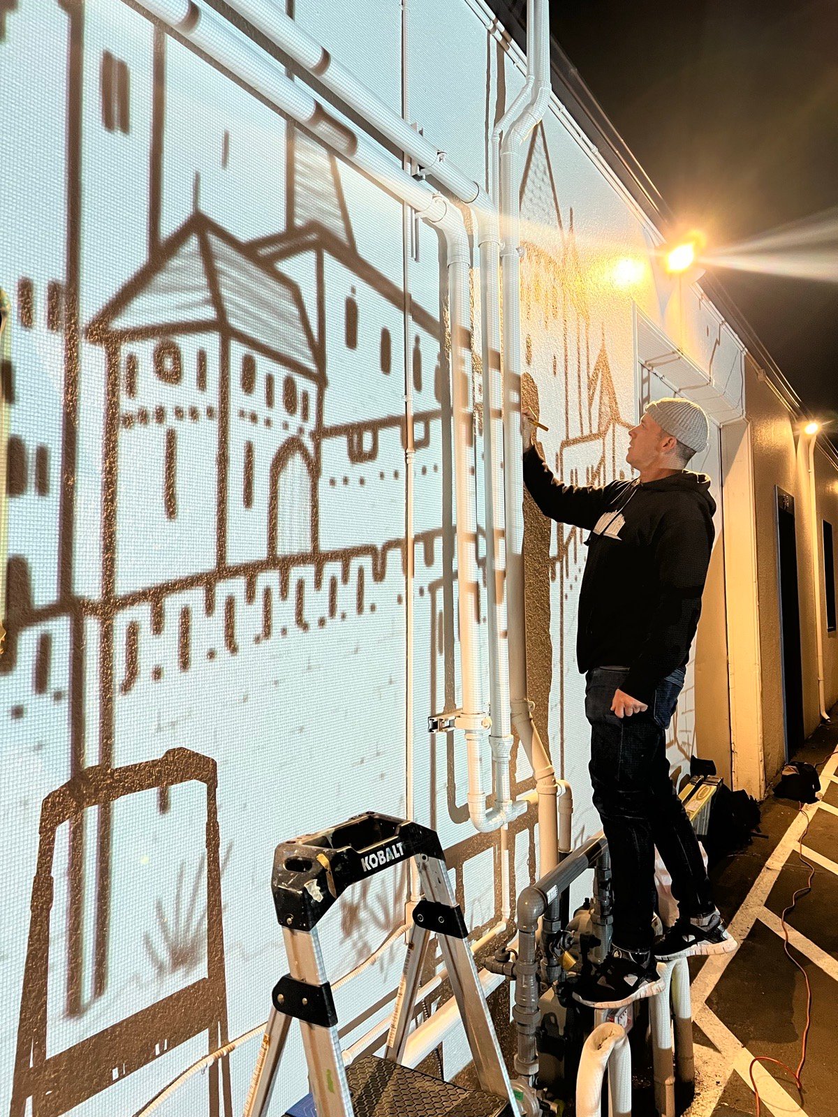 A muralist on a ladder at night, tracing the projected castle onto the wall